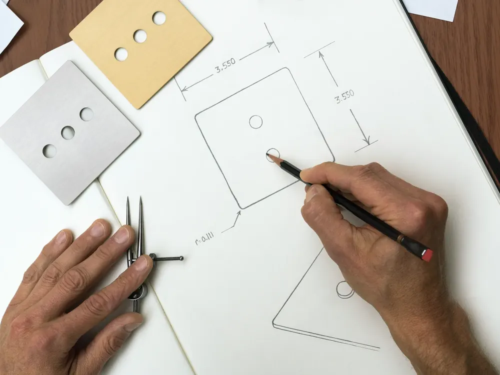 Designer sketching a keypad faceplate with dividers, metal faceplate samples beside the drawing