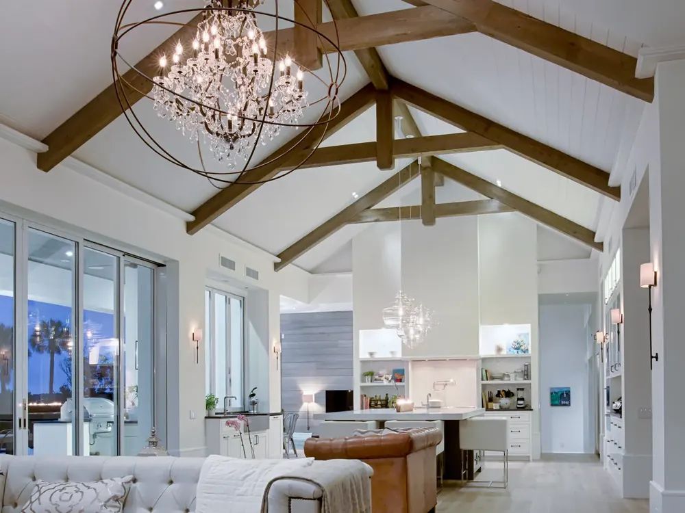 Open living room and kitchen at dusk under a vaulted beam ceiling, lit by a chandelier, wall sconces and pendants over the island